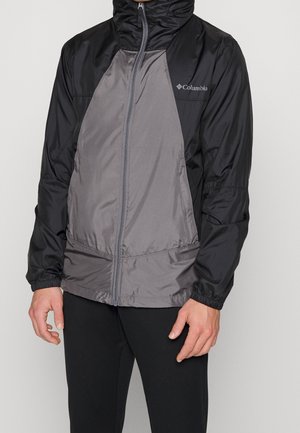 Man draagt een zwart-grijze Columbia zip-hoodie met elastische manchetten en een zwarte broek, staand tegen een effen achtergrond.