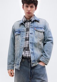 Νέος άνδρας που φοράει ανοιχτό μπλε τζιν μπουφάν πάνω από μαύρο και άσπρο καρό πουκάμισο, με μπλε τζιν παντελόνι, στέκεται μπροστά σε μια απλή φόντο.