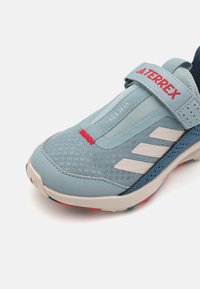Blue athletic shoe with mesh upper, suede accents, and red branding. Features a strap closure and textured sole for traction.