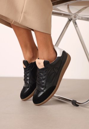 Persoon zittend op een stoel, draagt zwarte, beige en witte sneakers met gumzolen, beige broek boven de enkels cropped.