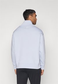 Lys blå genser med høy krage, med en subtil Nike-logo på baksiden. Glatt tekstur og ribbede mansjetter. Enkel design.