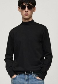 Cămașă neagră cu mâneci lungi și guler înalt, confecționată dintr-un material moale, asortată cu blugi albaștri și ochelari de soare negri. Design simplu, croi lejer.