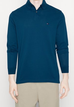Mand iført en langærmet navy blå polo-shirt med beige bukser, stående mod en ensfarvet hvid baggrund.