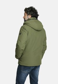 Chaqueta acolchada verde oliva con capucha, con una textura suave y detalles de costura. Usada sobre jeans azules, vista desde la parte posterior.