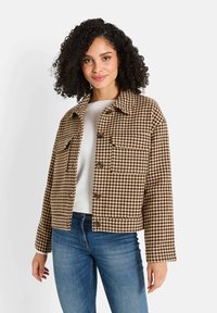 Veste en pied-de-poule marron avec un col, deux poches avant et des fermetures à bouton, portée avec un haut clair et un jean en denim.