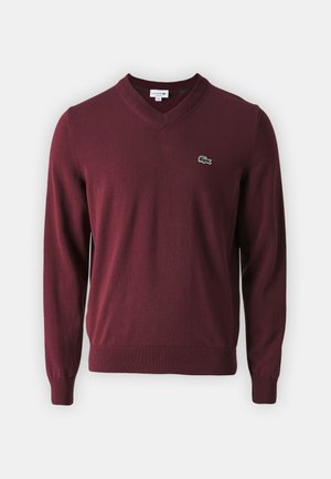 Burgundijas adīts džemperis ar V veida kakla izgriezumu, ribām pie piedurknēm un apakšmalas, ar mazu zaļu Lacoste krokodila logo kreisajā krūtī.