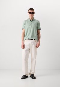 Světle zelené polo tričko s černými a bílými akcenty, spojené s volnými bílými kalhotami a černými nazouvacími botami. Minimalistický design.