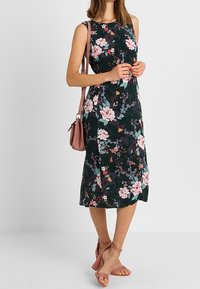 Abito floreale con sfondo scuro, senza maniche, al ginocchio, abbinato a una borsa rosa e sandali con cinturino coordinati.