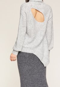 Hellgrauer Pullover mit hohem Kragen, offener Rückenschneider und schrägem Saum. Weiche Stricktextur, kombiniert mit einem dunkelgrauen Strickrock.