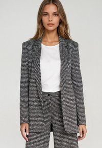Czarna i biała marynarka w pepitkę z konstrukcyjnymi ramionami. Gładki materiał, pojedyncze zapięcie na guzik i dwie przednie kieszenie. W zestawie z białą bluzką.