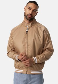 Chaqueta bomber color beige con cuello alto, cierre frontal y puños elásticos. Acentuada con rayas blancas en el dobladillo y los puños. Textura suave.