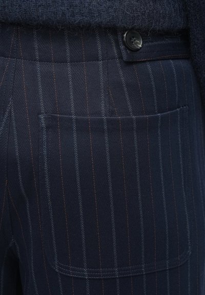 Marineblå bukser med et struktureret stof, der har smalle lodrette striber i blå og orange. Synlig baglomme og sort knapdetalje.