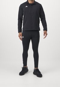 Czarna kurtka sportowa z zamkiem z przodu, z kontrastowym logo na piersi. Dopasowane czarne legginsy i czarne buty sportowe z sznurowadłami dopełniają stylizację.
