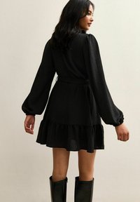 Vestido negro con mangas abullonadas largas, dobladillo con volantes y cintura ajustada. La tela parece ligera y texturizada, combinada con botas hasta la rodilla.