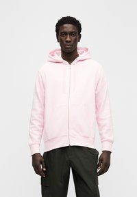 Różowa bluza z kapturem na zamek, wykonana z miękkiego materiału, z przednią kieszenią i kapturem na sznurek. Noszona z ciemnymi spodniami bojówkami. Białe logo na piersi.