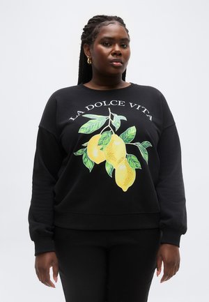 Naine punutud juustega, kellel on seljas must dressipluus, millel on sidrunite kujutis ja tekst "LA DOLCE VITA", ühevärvilisel heledal taustal.