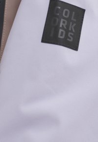 Vitt tyg med matt yta som har en svart rektangulär etikett präglad med texten "COLOR KIDS." Inga ytterligare mönster eller dekorationer.