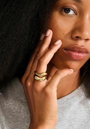 Gold metal rings with a smooth, wavy design worn on a finger, alongside a hand resting against the face. Skin has a natural finish.