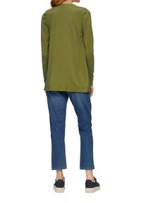 Olijfgroen top met lange mouwen en een ontspannen pasvorm, met zijsplitten. Gecombineerd met blauwe denim jeans en zwarte plateausneakers.
