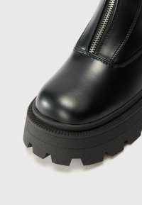 Botte de cheville en cuir noir avec semelle plateforme épaisse, dotée d'un bout arrondi et de détails en fermeture éclair argentée sur le côté. Semelle extérieure texturée pour une meilleure adhérence.