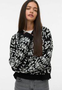 Giovane donna con lunghi capelli castani che indossa un maglione a righe bianco e nero e jeans grigi, in posa contro uno sfondo semplice.