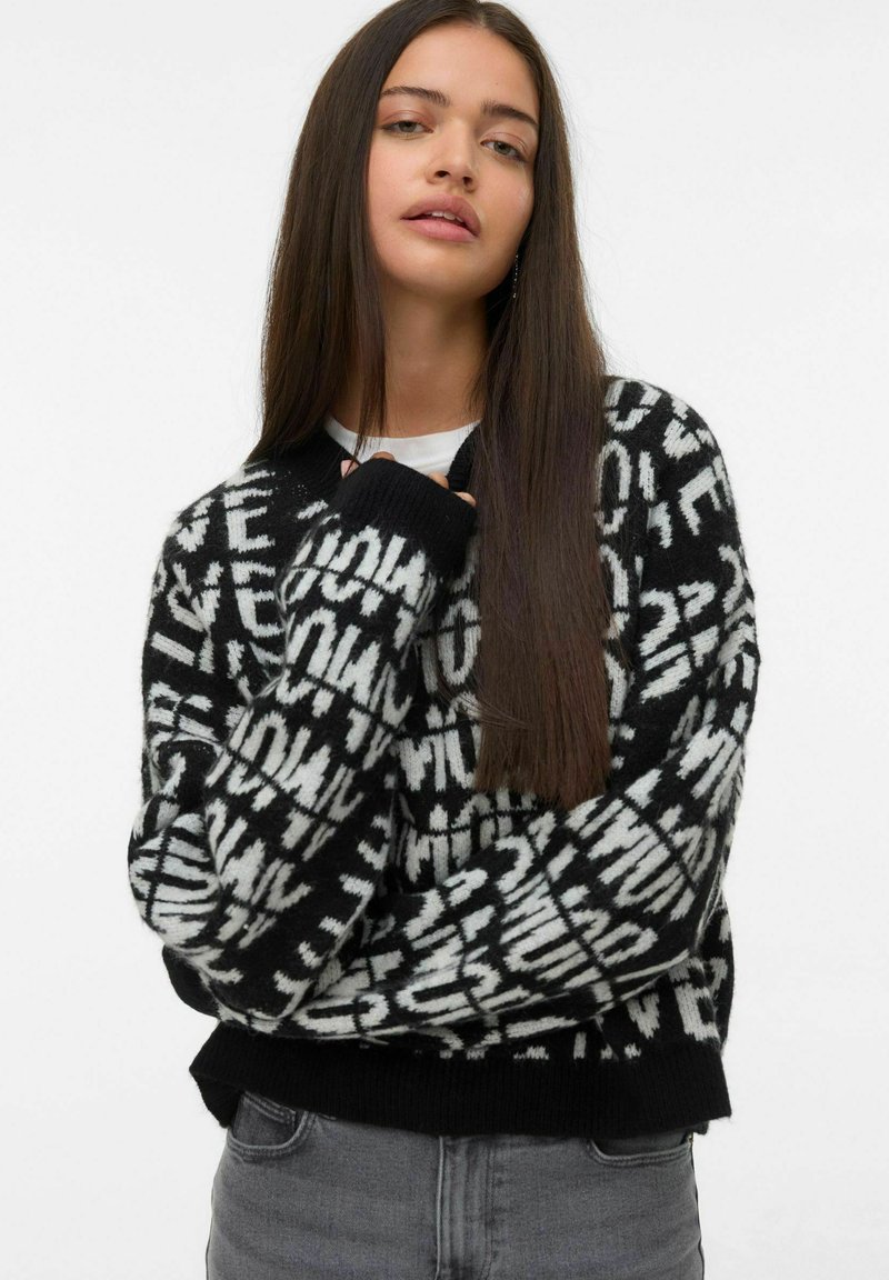 Giovane donna con lunghi capelli castani che indossa un maglione a righe bianco e nero e jeans grigi, in posa contro uno sfondo semplice.