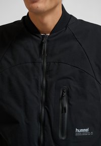 Schwarze Bomberjacke mit durchgehendem Reißverschluss, geripptem Kragen und strukturiertem Stoff. Verfügt über eine Brusttasche mit verdecktem Reißverschluss und Branding.