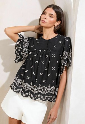 Blusa nera con raffinati ricami bianchi e orlo a scallop, caratterizzata da dettagli plissettati e maniche a campana. Indossata con pantaloni corti bianchi.