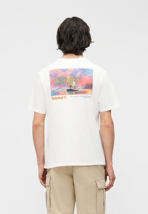 Inimene, kes kannab valget Timberlandi T-särki, millel on seljal värvikas vanakooliline trükk kahest inimesest, kes kalastavad jõe ääres.