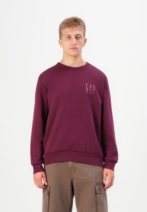 Maronfarvet sweatshirt med ribbet rund halsudskæring, lange ærmer og 'GAP' logo i lyse lilla på venstre bryst. Materialet er en blanding af bomuld med en glat tekstur.