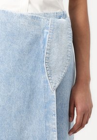 Denimskjørt i lyseblått med en framlomme med spiss form. Tøyet har en falmet tekstur med synlige sømmer langs kantene.