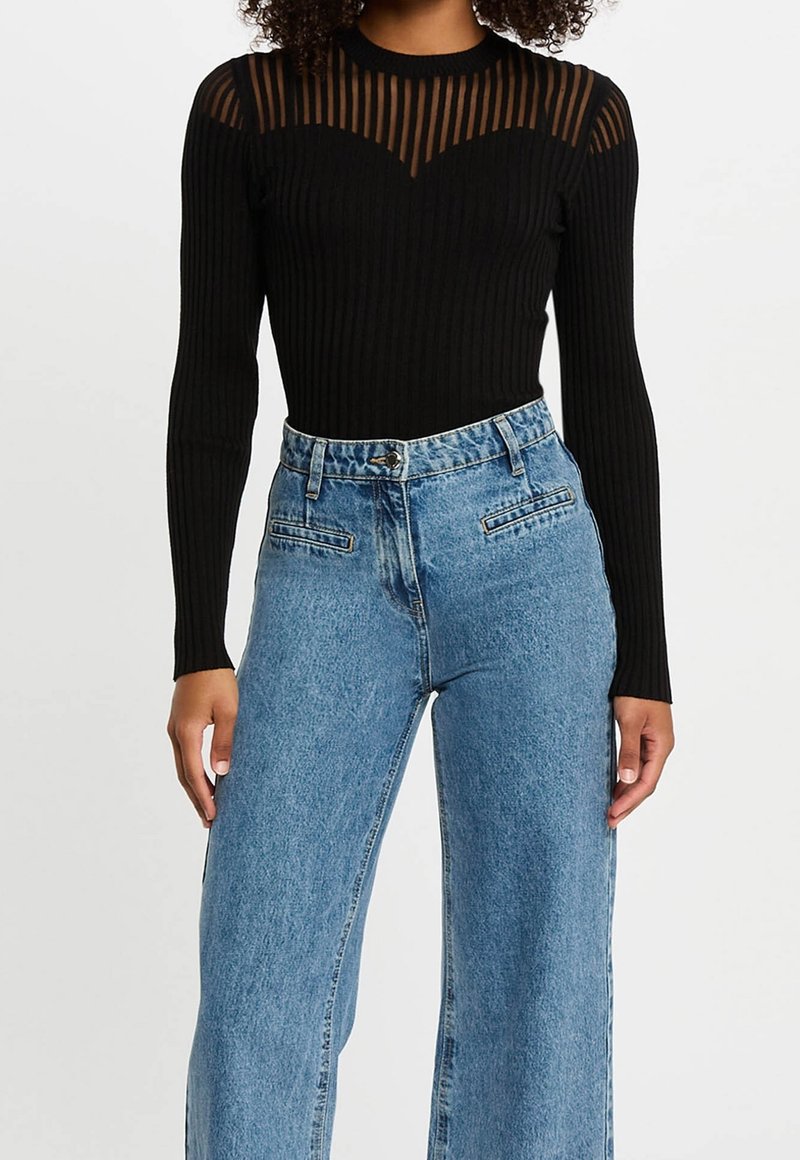 Femme portant un haut noir côtelé à manches longues avec un empiècement transparent rayé et un jean bleu ample taille haute sur un fond uni.