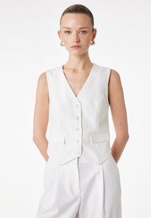 Donna con capelli pettinati all'indietro che indossa orecchini a cerchio dorati, un gilet bianco senza maniche con bottoni e pantaloni bianchi abbinati, in piedi davanti a uno sfondo bianco.