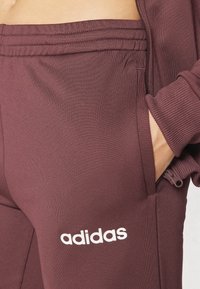 Maroon athletic pants made of soft fabric, featuring a side pocket, elastic waistband, and white "adidas" logo on the thigh.