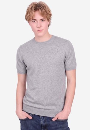 Giovane uomo con capelli chiari che indossa un maglione grigio a maniche corte e jeans blu, in piedi con le mani in tasca davanti a uno sfondo semplice.