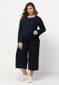 Top blu navy a maniche lunghe con dettaglio annodato davanti, abbinato a culotte nere con pois bianchi e sneakers bianche. Sfondo semplice.
