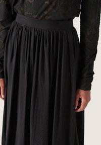 Gonna nera plissè con una superficie liscia, ampia cintura elastica e dettagli in pizzo floreale intricato su una blusa a maniche lunghe trasparente.
