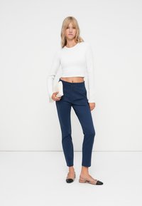 Hvit langermet cropped topp kombinert med mørkeblå skreddersydde bukser, iført beige flate sko med svart tåhette, minimalistisk clutch holdt i hånden.