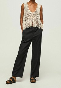 Szydełkowa kamizelka w kolorze kremowym z frędzlami, zestawiona z luźnymi czarnymi spodniami oraz czarnymi sandałami z akcentami klamry.