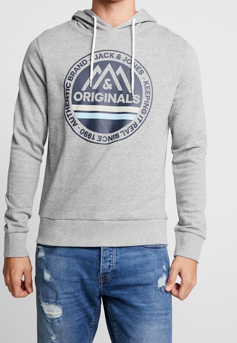 Homme portant un sweat à capuche gris clair avec le logo Jack & Jones Originals et un jean bleu délavé, debout devant un fond uni.