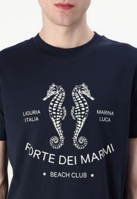Jūras zila kokvilnas T-kreklu ar baltajiem jūraszirdziņu motīviem, tekstu, kas ietver "Forte dei Marmi", un ribinātu apkakli.