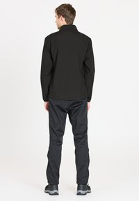 Musta vedenkestävä takki korkealla kauluksella, yhdistettynä tummanharmaisiin hoseihin. Takissa on sileä pinta ja säädettävät hihansuut.