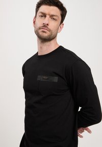 Czarna koszulka z długim rękawem wykonana z bawełny, posiadająca małą kieszonkę na piersi z prostokątną metką. Prosty, dopasowany krój z okrągłym dekoltem.