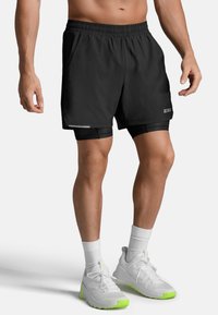 Mannelijk model gekleed in zwarte sportshorts met dubbele laag en witte sneakers met neon groene zolen tegen een effen achtergrond.