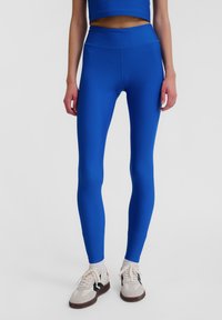 Blå leggings med hög midjeresår, tillverkade av ett stretchigt material. De har en slät yta och en figurnära design, kombinerade med avslappnade sneakers.