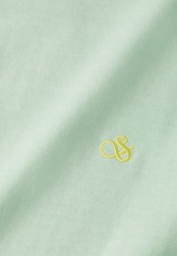 Tejido de un verde menta claro con una textura suave, que presenta un pequeño logo bordado en amarillo con la inscripción "as" en la esquina inferior derecha.