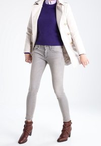 Persona con un cappotto bianco, un maglione viola, jeans skinny grigio chiaro e stivali con tacco marroni, in piedi con le gambe divaricate su uno sfondo bianco.