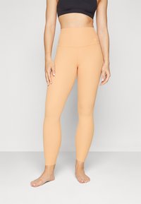 Leggings color pesco con vita alta, texture a costine e design aderente. Accento del logo in vita. Modella a piedi nudi in posizione eretta.