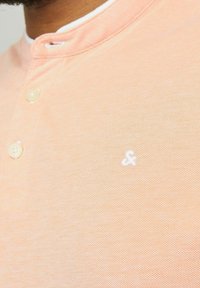 Breskva polo majica s teksturiranim materijalom, koja sadrži bijeli izvezeni logo i tri dugmeta na ovratniku.
