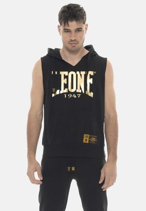 Giovane uomo con felpa nera senza maniche e pantaloni con logo dorato "LEONE 1947", in piedi davanti a uno sfondo bianco uniforme.